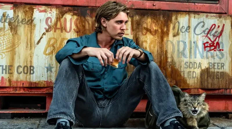 Austin Butler como Hank sentado en el suelo con un gato atigrado a su lado frente a una pared oxidada con grafitis en Atrapado Robando.