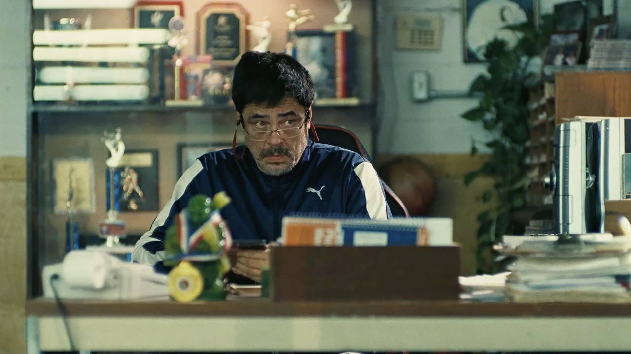 Benicio del Toro como el Sensei Sergio sentado en su escritorio rodeado de trofeos en una escena de Una batalla tras otra.
